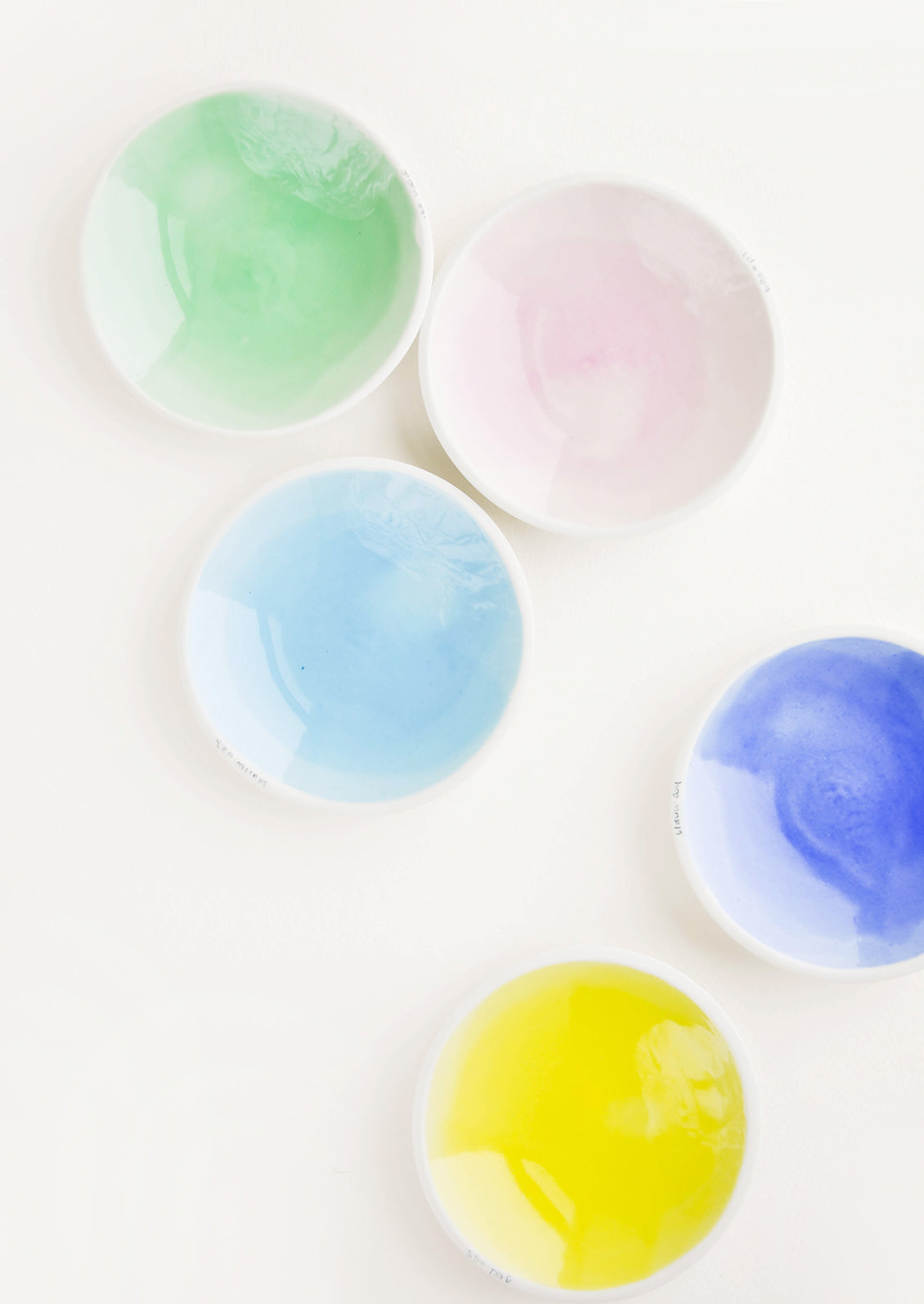 Five shallow porcelain dishes in light blue, blue, pale pink, green, and yellow with shiny finishes and matte white rims.