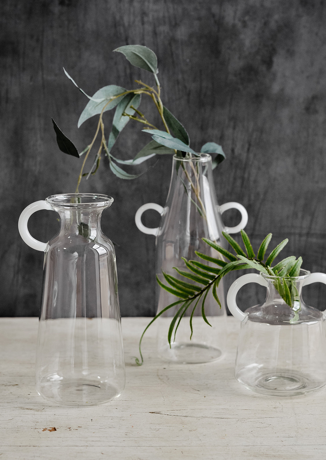 A trio of glass vases with white curved handles, displaying greenery.