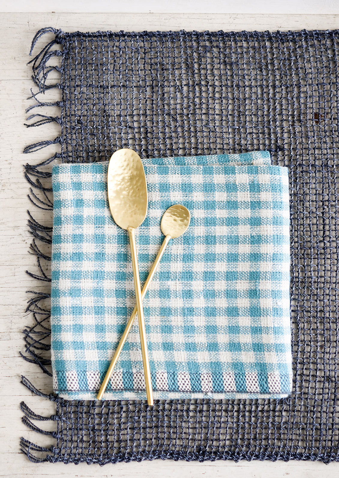 A blue-grey woven straw placemat with gingham napkin and gold spoons.