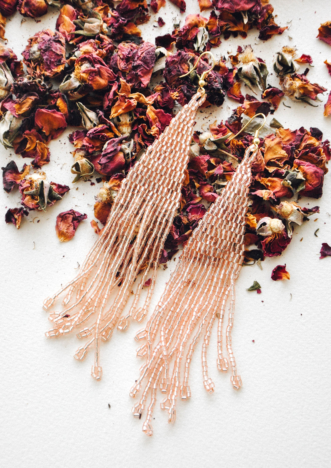 Light pink iridescent beaded fringe earrings rest atop a bed of dried rose petals.